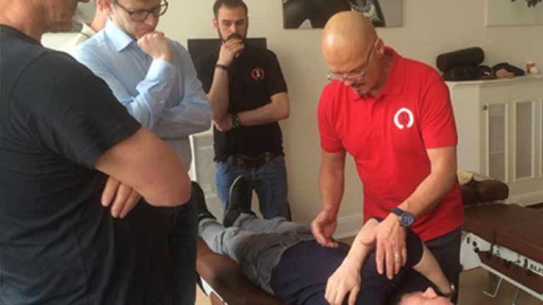 Chiropraktik Campus Kiseo Vier Männer stehen um einen liegenden Mann herum, drei sind eher beobachtend, ein Mann mit rotem T-Shirt behandelt die liegende Person