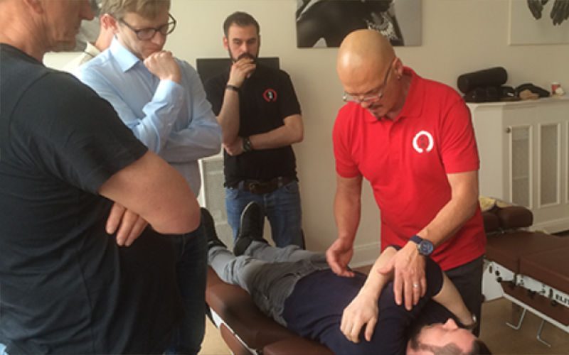 Chiropraktik Campus Kiseo Vier Männer stehen um einen liegenden Mann herum, drei sind eher beobachtend, ein Mann mit rotem T-Shirt behandelt die liegende Person