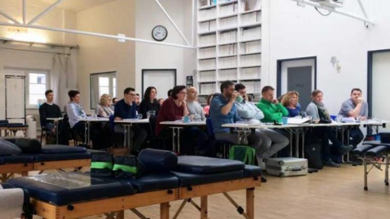 Chiropraktik Campus Eine Unterrichtssituation mit mehreren an Tischen sitzenden Personen in einem großen Raum, vor den Personen stehen mehrere Liegen.