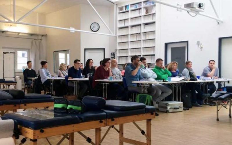 Chiropraktik Campus Eine Unterrichtssituation mit mehreren an Tischen sitzenden Personen in einem großen Raum, vor den Personen stehen mehrere Liegen.