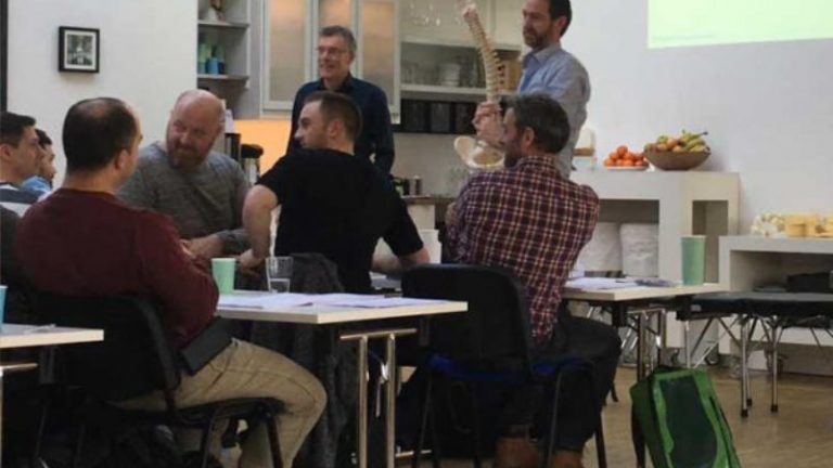 Chiropraktik Campus Einige Männer sitzen an Tischen, zwei Männer stehen vor ihnen, einer hält ein Modell einer Wirbelsäule in der Hand