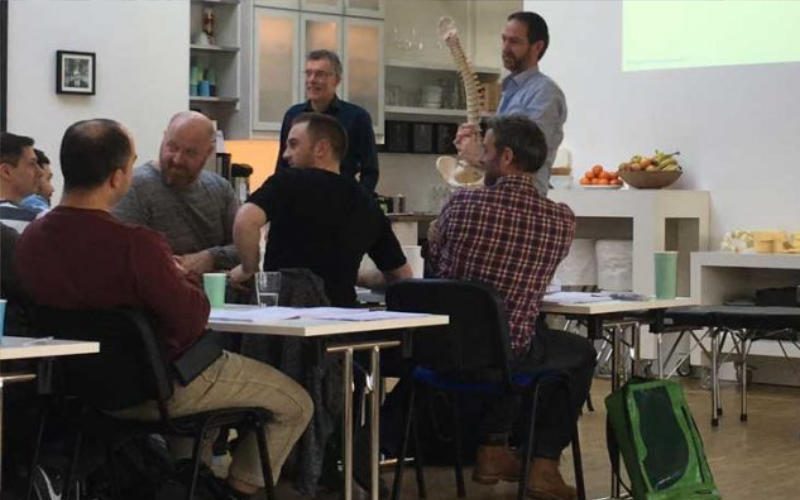 Chiropraktik Campus Einige Männer sitzen an Tischen, zwei Männer stehen vor ihnen, einer hält ein Modell einer Wirbelsäule in der Hand