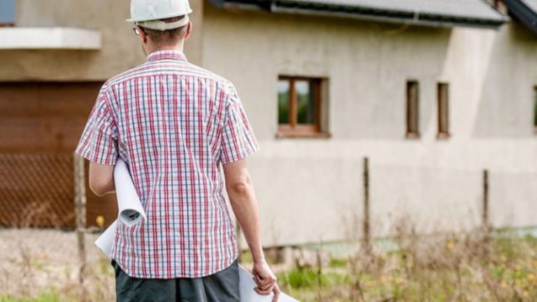 Mann mit Bauhelm und Bauzeichnugen unter dem Arm vor Haus