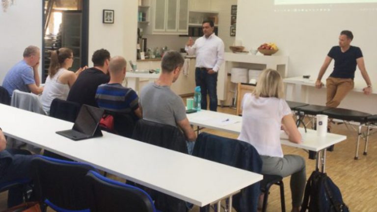 Chiropraktik Campus Studium Einige Personen sitzen an Tischen, zwei Männer stehen vor ihnen und erklären etwas