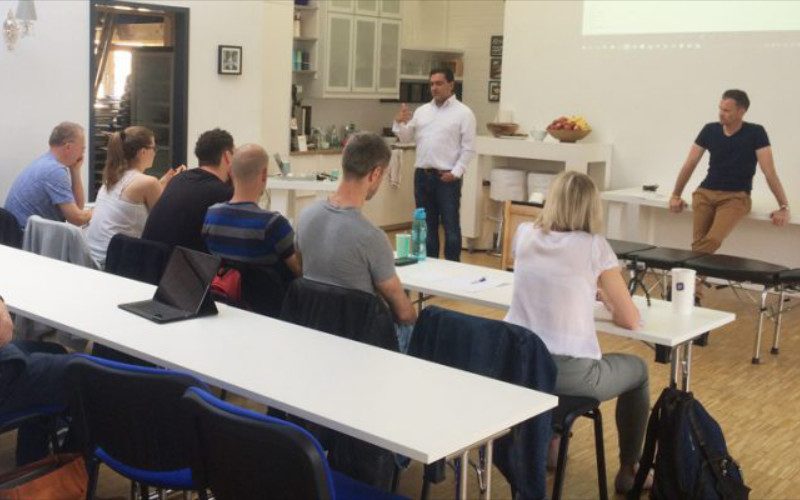 Chiropraktik Campus Studium Einige Personen sitzen an Tischen, zwei Männer stehen vor ihnen und erklären etwas