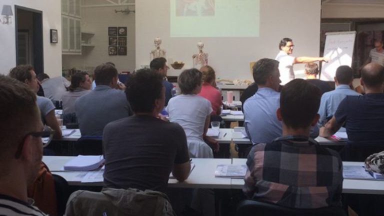 Chiropraktik Campus Unterricht In einem Seminarraum sitzen einige Personen an Tischen, vor ihnen steht ein Mann und erklärt einige Inhalte