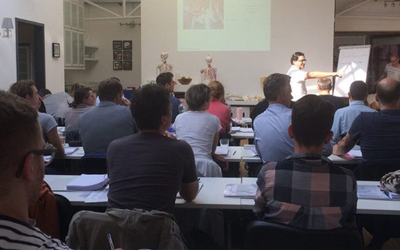 Chiropraktik Campus Unterricht In einem Seminarraum sitzen einige Personen an Tischen, vor ihnen steht ein Mann und erklärt einige Inhalte