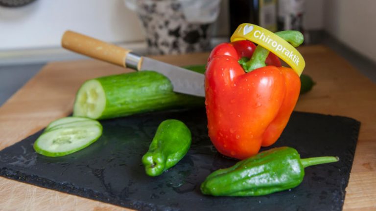 Auf einem Schieferbrett liegt eine Gurke, 2 grüne Paprikas und eine rote Paprika, auf dieser liegt ein Gummiarmband mit der Aufschrift: l love Chiropraktik