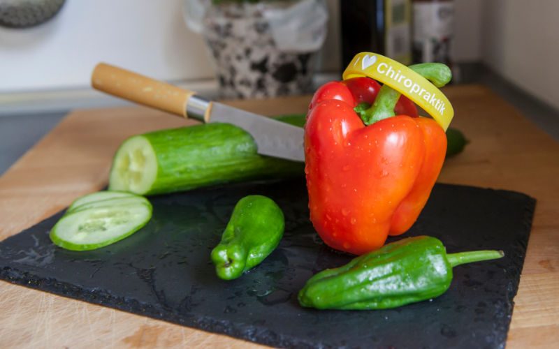 Gesunde Ernährung Auf einem Schieferbrett liegt eine Gurke, 2 grüne Paprikas und eine rote Paprika, auf dieser liegt ein Gummiarmband mit der Aufschrift: l love Chiropraktik