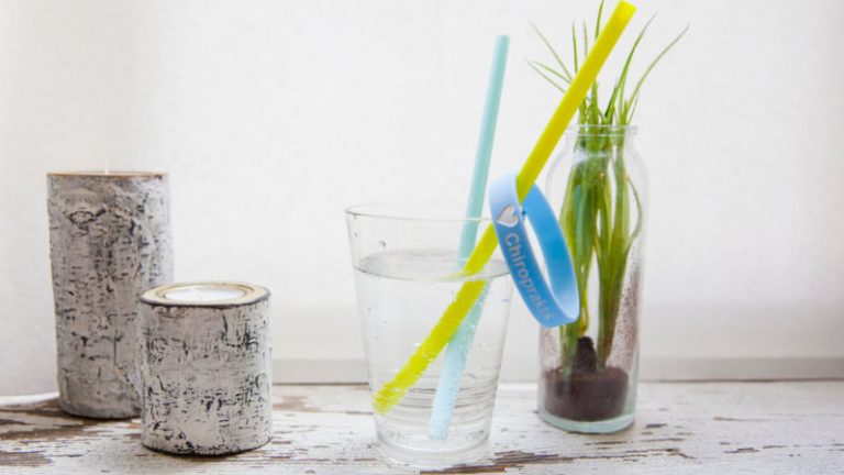 Zwei Kerzen stehen neben einem Glas das mit Wasser gefüllt ist und in dem zwei Strohhalme stecken, an einem gelben Strohhalm hängt ein blaues Gummiarmband, im Hintergrund steht eine Pflanze