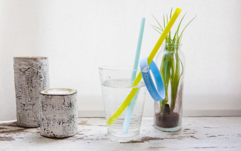 Trinken Zwei Kerzen stehen neben einem Glas das mit Wasser gefüllt ist und in dem zwei Strohhalme stecken, an einem gelben Strohhalm hängt ein blaues Gummiarmband, im Hintergrund steht eine Pflanze