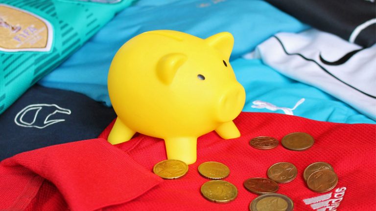 Sparschwein Ein gelbes Sparschwein steht auf verschiedenen bunten Trikots, vor diesem liegen einige Euromünzen