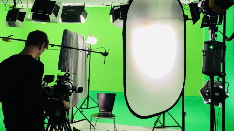 Studio Grün Hamburg Ein Filmstudio mit einem Green Screen und Filmlicht, Scheinwerfern und Abblenden, eine Person steht hinter einer Kamera die auf einen leeren Stuhl gerichtet ist.