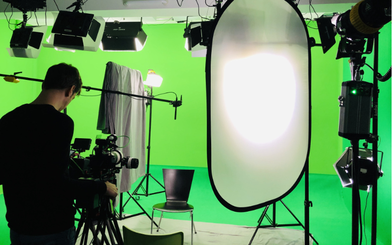 Ein Filmstudio mit einem Green Screen und Filmlicht, Scheinwerfern und Abblenden, eine Person steht hinter einer Kamera die auf einen leeren Stuhl gerichtet ist.