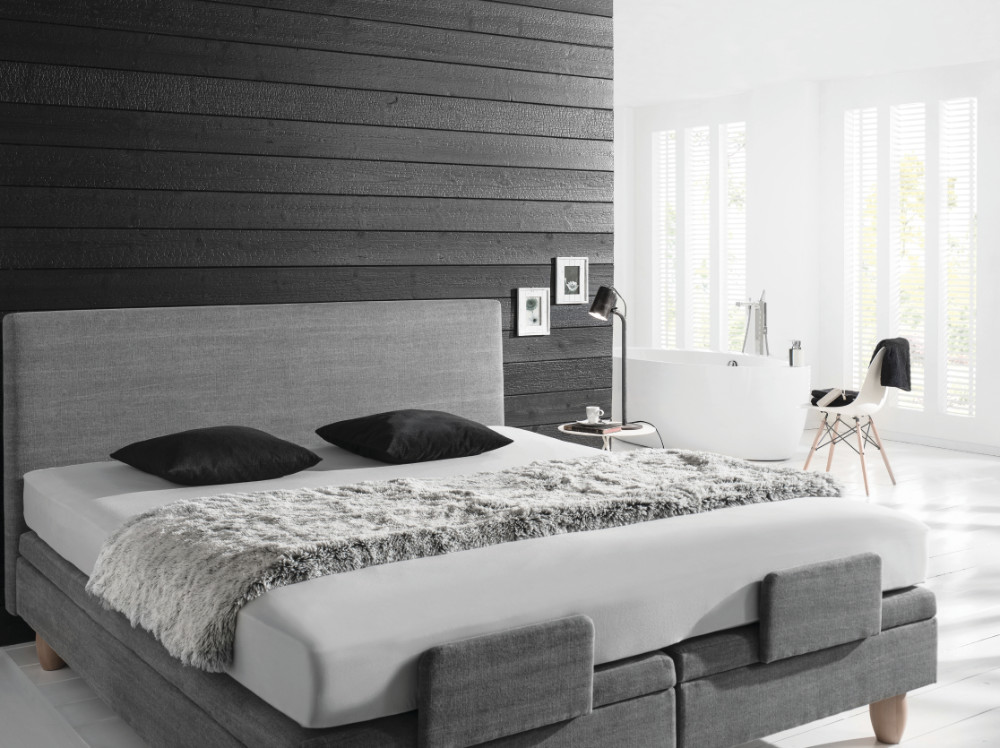 Ein großes Doppelbett mit 2 Kissen und einer Felldecke steht vor einer Holzwand, im Hintergrund steht eine ovale Badewanne und ein Stuhl.
