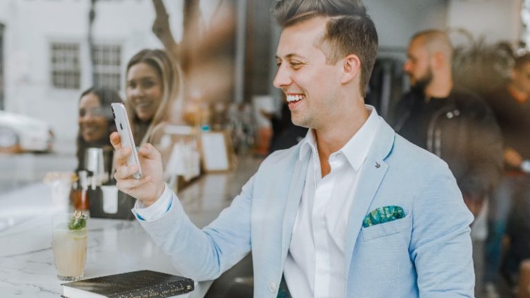Ein chic gekleideter junger Mann in grauem Jacket, sitzt an einem Bartisch und lächelt sein Handy an. Hinter ihm stehen weitere Menschen die ebenfalls lächeln.