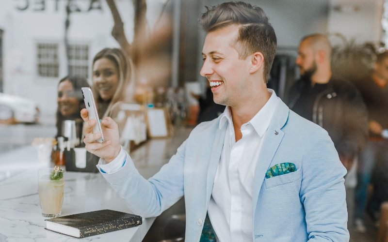 Lächelnder Mann Ein chic gekleideter junger Mann in grauem Jacket, sitzt an einem Bartisch und lächelt sein Handy an. Hinter ihm stehen weitere Menschen die ebenfalls lächeln.