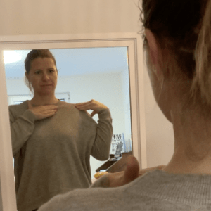 Eine Frau steht vor einem Spiegel und berührt ihre rechte Schulter mit beiden Händen.