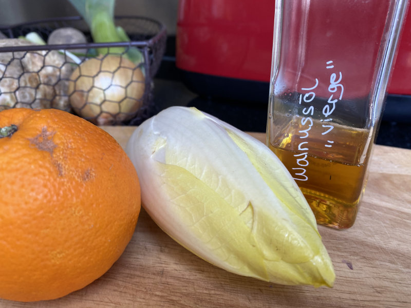 Eine Orange, ein Chicoree und Ein Glass mit Walnussöl stehen auf einem Holztisch.