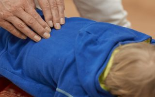 FriedemannTheill_Chiropraktik bei Kindern_Generation Lockdonw-NL Hände eines Chiroprakters bei der Justierung von einem Kind in blauem Hoody.