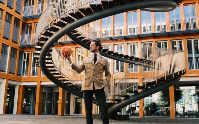 The Suit League Ein Mann im beigen Jacke steht vor einer spiralen Treppe und hält ein Kübis in der Hand.