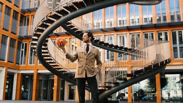 Ein Mann im beigen Jacke steht vor einer spiralen Treppe und hält ein Kübis in der Hand.