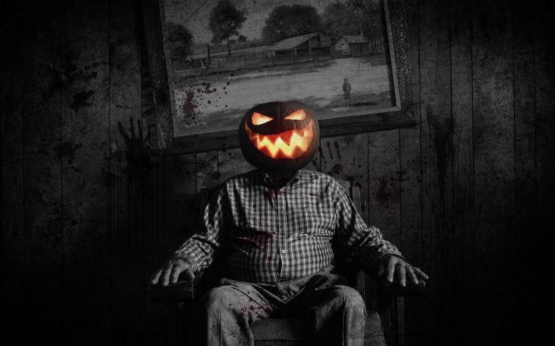 Halloween Schwarz-weisses Halloween Foto mit einem Mann der auf einem Sessel sitzt und als Kopf ein leuchtendes Halloween Kürbis hat.