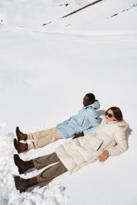 Eine Frau in weissen Mantel und ein Man in hell blauer Jacke liegen im Schnee nebeneinander und tragen Sonnenbrille.