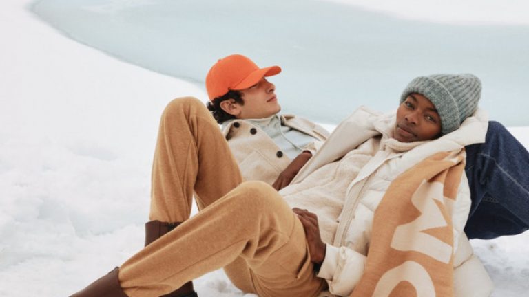 Ein Man mit orangenen Cappy und eine Frau mit grauer Mütze und Tom Tailor Schall liegen im Schnee.