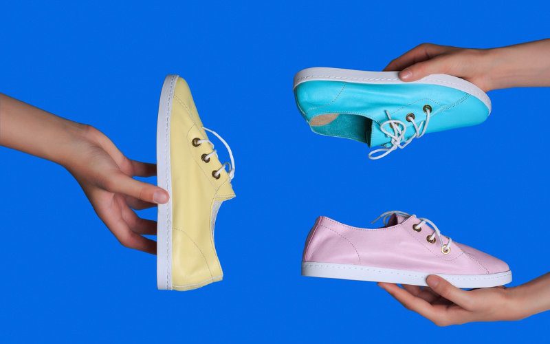 Schuhe Drei Hände halten jeweils drei Turnschuhe in Blau, Pink und Gelb vor einem blauen Hintergrund.