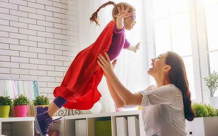 Mama wirft ihre kleine Tochter im Superhelden Kostümspielerisch in die Luft