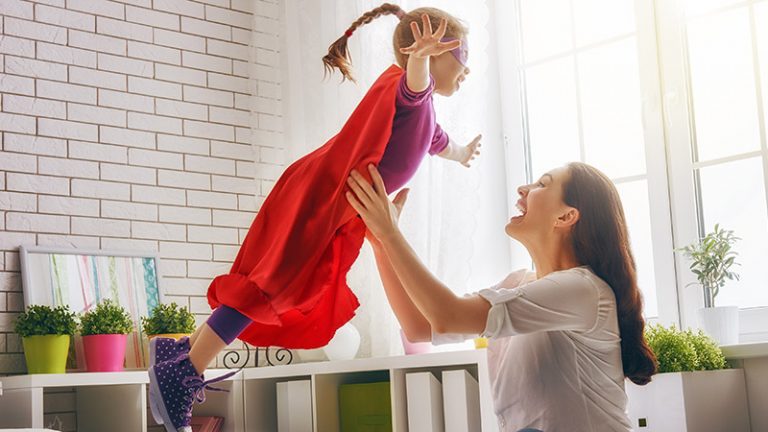 Mama wirft ihre kleine Tochter im Superhelden Kostümspielerisch in die Luft
