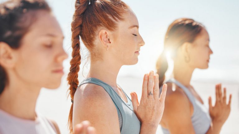 Yoga Meditation 3 Frauen haben die Hände vor der Brust zusammengelegt und geschlossene Augen. Sie tragen sportliche Kleidung und im Hintergrund scheint die Sonne.
