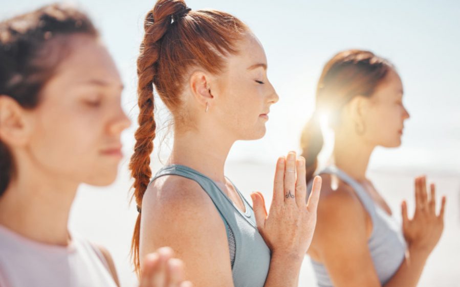 Yoga Meditation 3 Frauen haben die Hände vor der Brust zusammengelegt und geschlossene Augen. Sie tragen sportliche Kleidung und im Hintergrund scheint die Sonne.