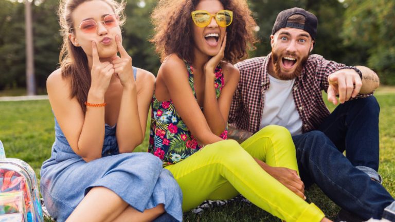 Drei glückliche junge Personen sitzen auf einer Wiese und machen lustige Gesichter.