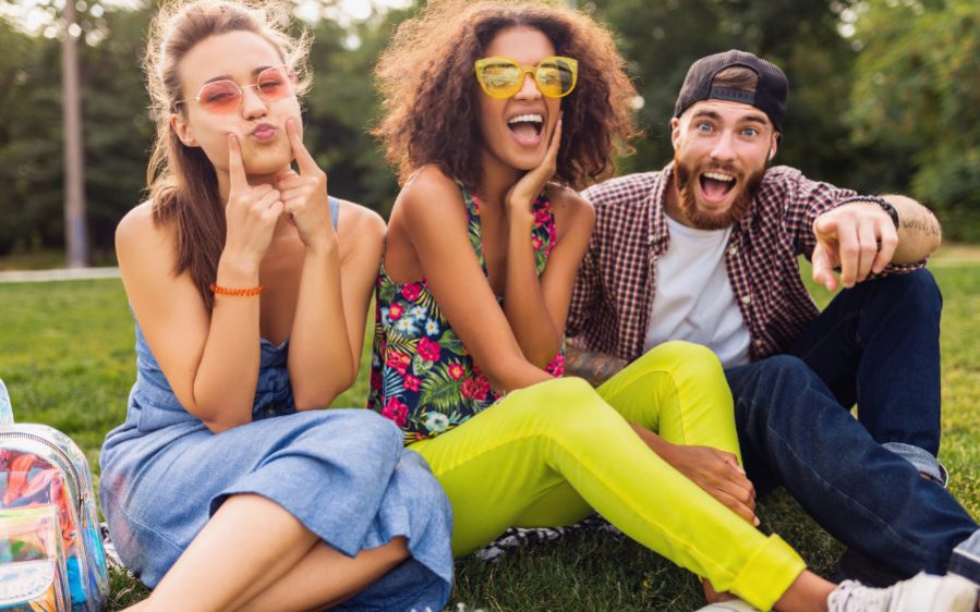 Drei glückliche junge Personen sitzen auf einer Wiese und machen lustige Gesichter.