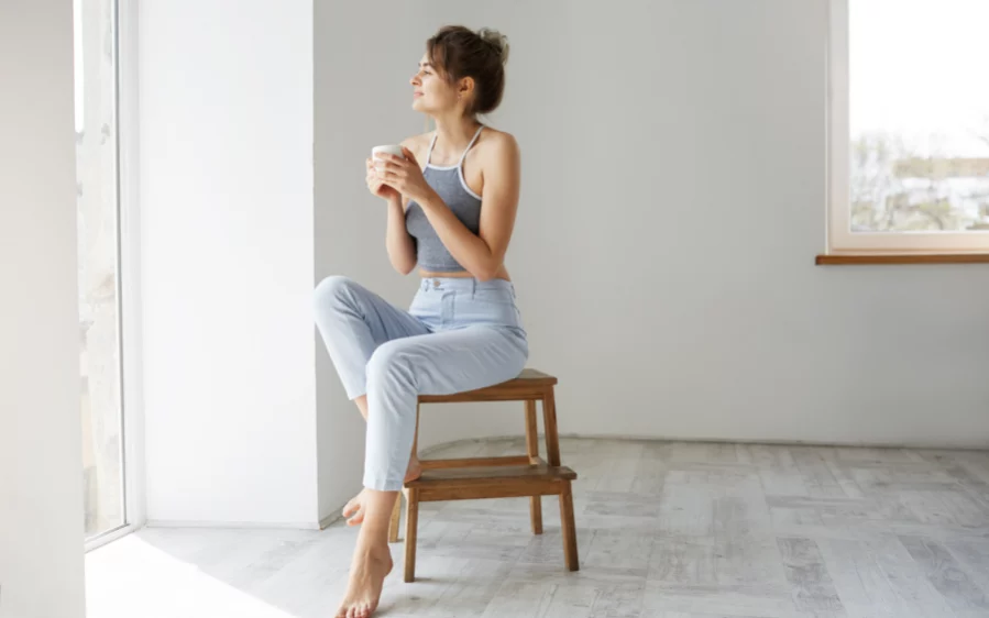 junge-bruenette-frau-laechelnd-die-tasse-betrachtet-die-fenster-sitzt-auf-stuhl-am-morgen_900x600px Junge Frau sitzt am Fenster auf einem Hocker, genießt ihren Kaffee und schaut lächelnd nach draußen.