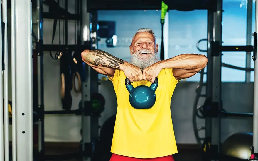 Älterer Mann in gelbem T-Shirt und mit weißen Haaren und Bart trainiert im Fitnessstudio. Er hält in seinen Händen ein Gewicht.