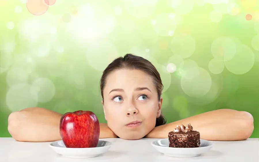 attraktive-junge-frau-trifft-ihre-wahl-zwischen-apfel-und-kuchen-gesundes-lifestyle-konzept_900x600 Frau die sich auf einen Tisch stützt und nachdenklich in die Luft schaut. Vor ihr befindet sich ein Apfel und ein Stück Kuchen