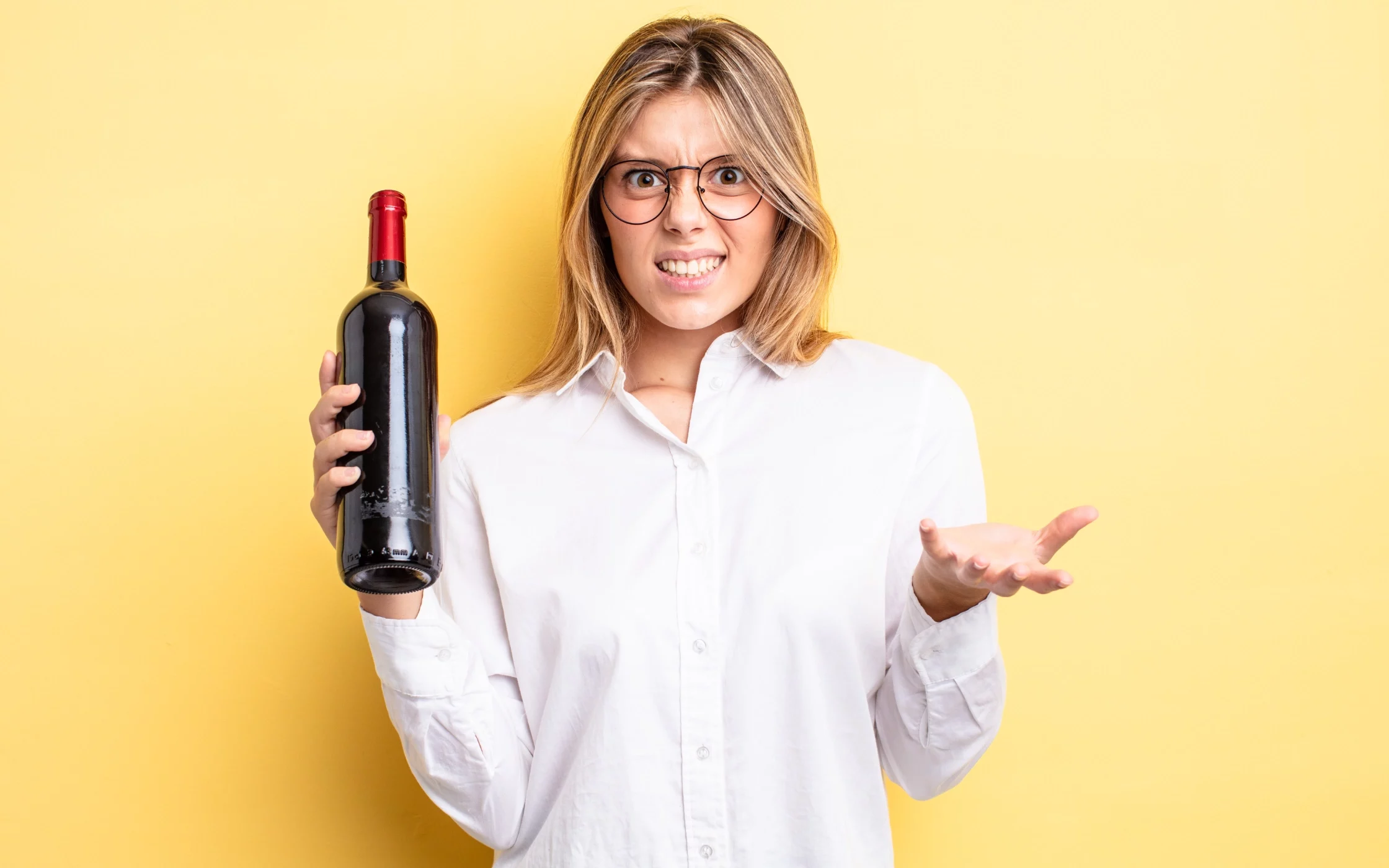 Alkohol_2400x1400 Eine blonde Frau, die ein weißes Hemd und eine Brille trägt, hält eine Rotweinflasche in der Hand und guckt den Beobachter skeptisch an.