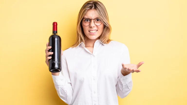 Eine blonde Frau, die ein weißes Hemd und eine Brille trägt, hält eine Rotweinflasche in der Hand und guckt den Beobachter skeptisch an.