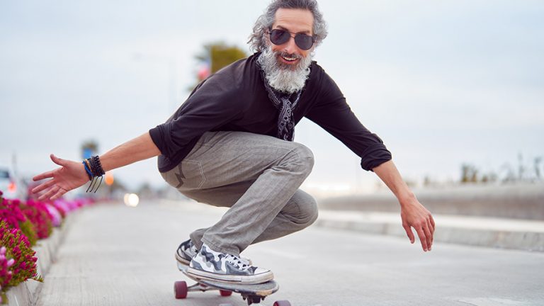 mature man white beard skateboarding on street and having fun Alter Mann der in coolen Klamotten Skateboard fährt